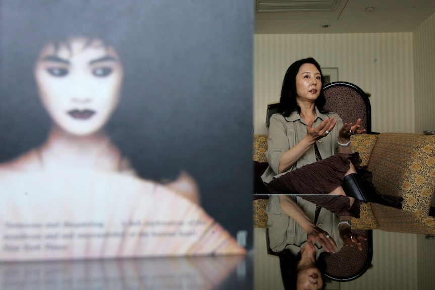 A woman, in focus, sits well behind a blurred book cover as she is interviewed.  She has her hands outstretched, palms upwards