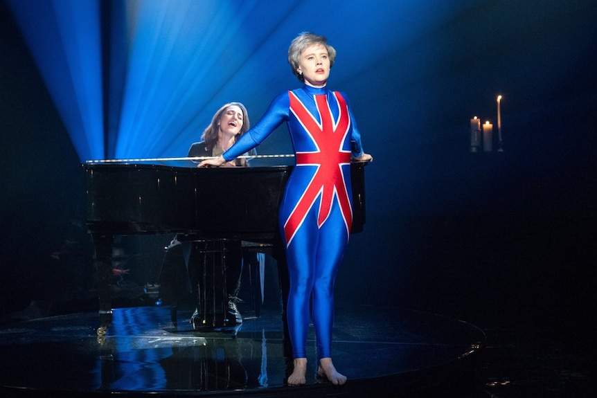 Kate McKinnon wearing a cropped grey wig and Union Jack unitard while Sara Bareilles sings and plays piano.