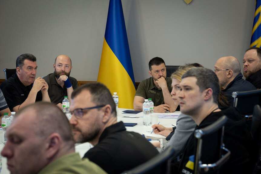 Volodymyr Zelenskyy holds an emergency meeting with people in civilian clothes seated around a table with a Ukraine flag behind.