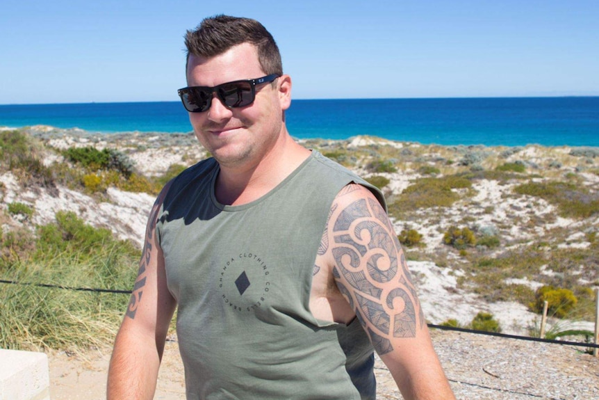 A man wearing a singlet and sunglasses walking along a beach.