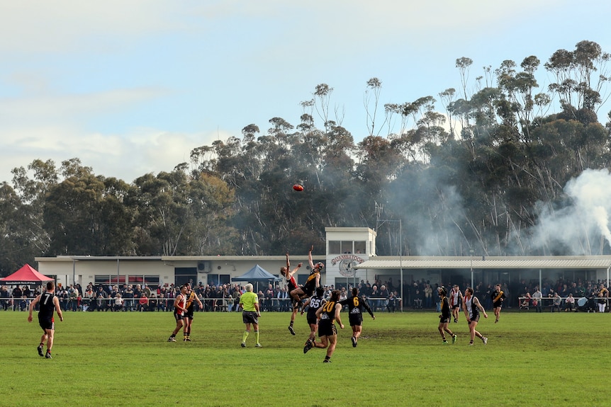 Two players jump for a ball in the middle of a country football ground, watched on by other players and a row of spectators