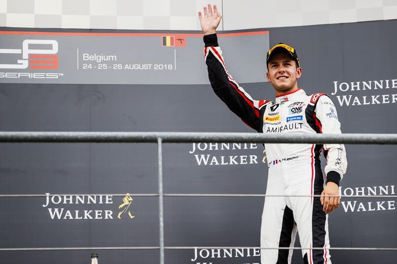 A man in a white racing suit stands on a podium while waving