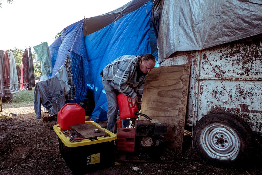 A man pours petrol into a generator near a tarp covered caravan.