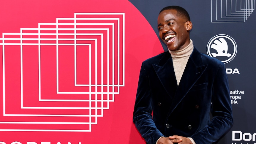 Ncuti Gatwa smiles in a velvet suit in front of a poster for the European film awards.