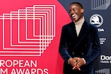 Ncuti Gatwa smiles in a velvet suit in front of a poster for the European film awards.