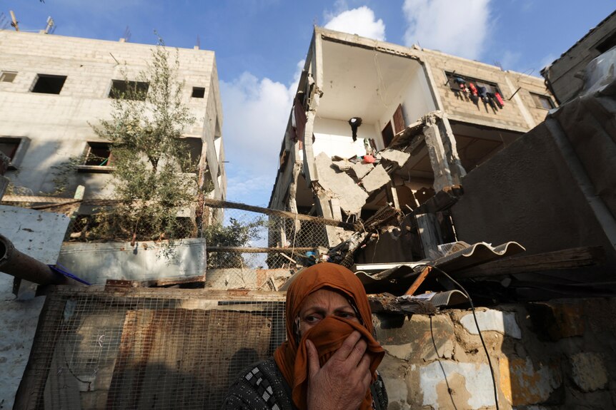 A woman in an orange head scarf appers distressed in front of a damaged house. 