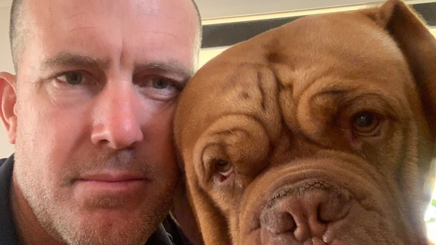 A selfie of a serious man with shaved hair next to his large brown dog.