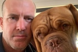 A selfie of a serious man with shaved hair next to his large brown dog.