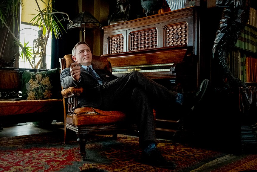 Daniel Craig wears full suit and reclines in arm chair next to upright piano in ornately decorated interior.