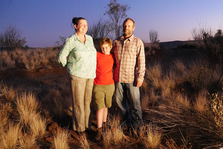 Boy in red top stands in middle of man and woman with bushland behind them.