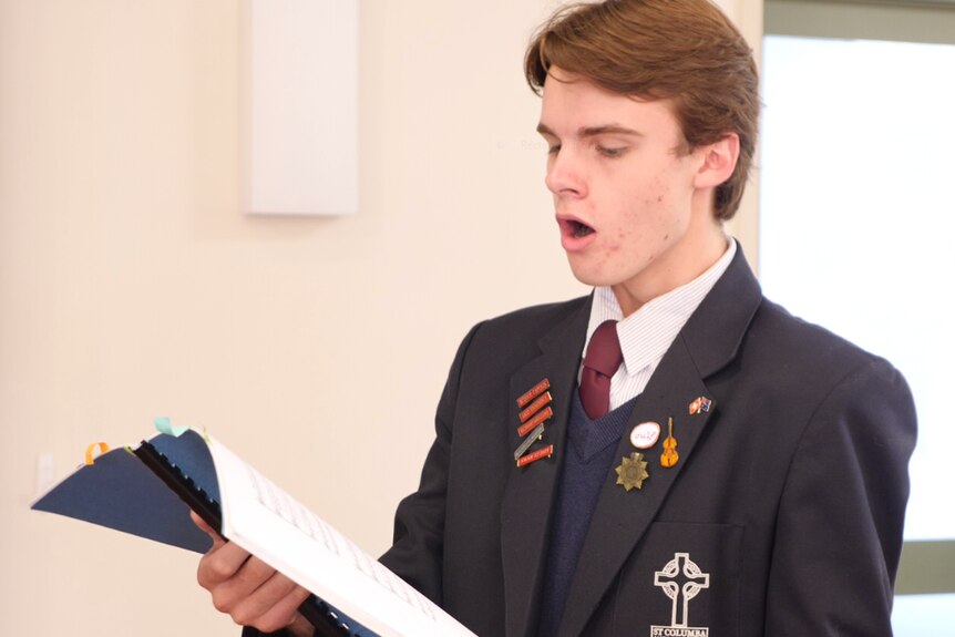 A teenage boy in a school blazer singing from a book