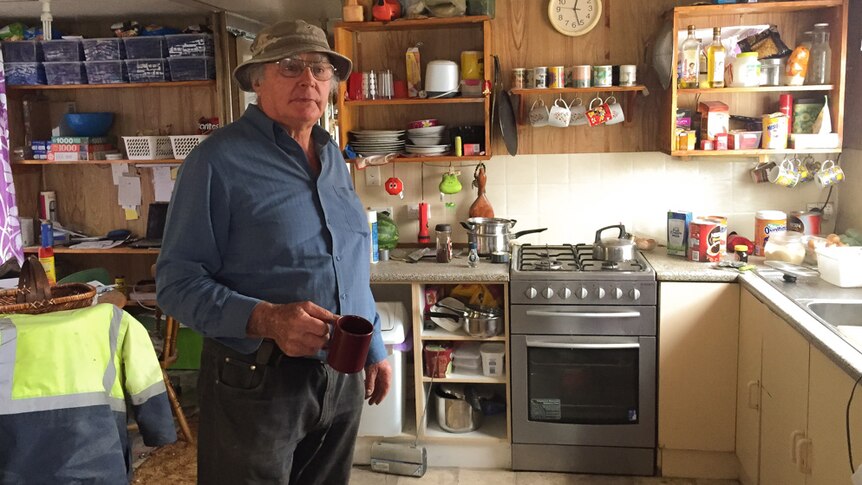 Rodney West holding a mug while standing in the kitchen of his houseboat, which is crammed with household belongings.