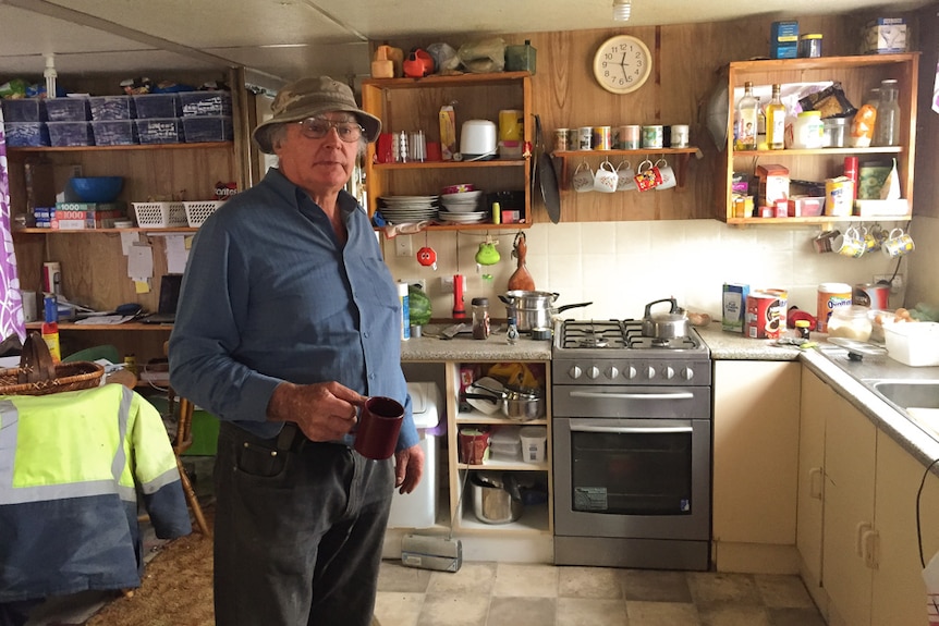 Rodney West holding a mug while standing in the kitchen of his houseboat, which is crammed with household belongings.