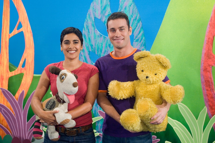 A woman and man stand next to each other holding toys with a colourful background. 