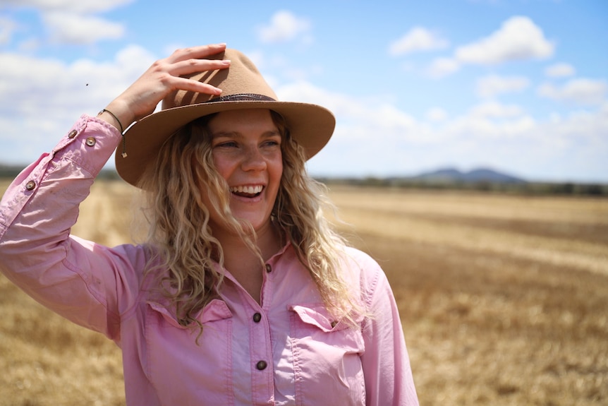 A young woman with shoulder-length, wavy blonde hair wears a pint work shirt and an Akubra hat