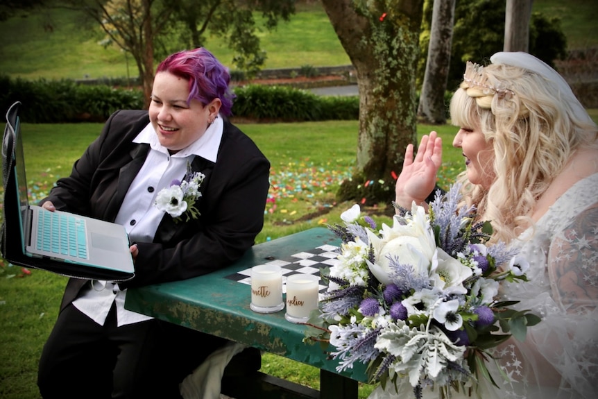 Jessica King in a black suit and Emily Sinclair in a white bridal gown adorned with stars smile and wave at a laptop.
