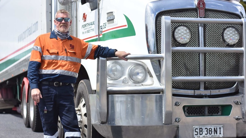 A man in a high vis orange work shirt is dwarfed by a big white freight truck branded "Porthaul"