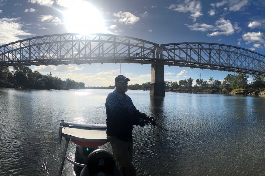 A man stands up in a boat while fishing in a river with a bridge behind him on a sunny day