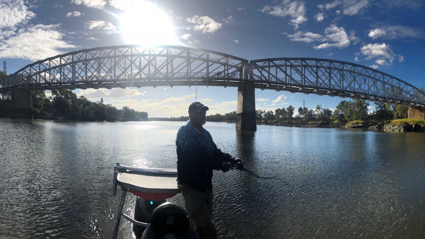 A man stands up in a boat while fishing in a river with a bridge behind him on a sunny day