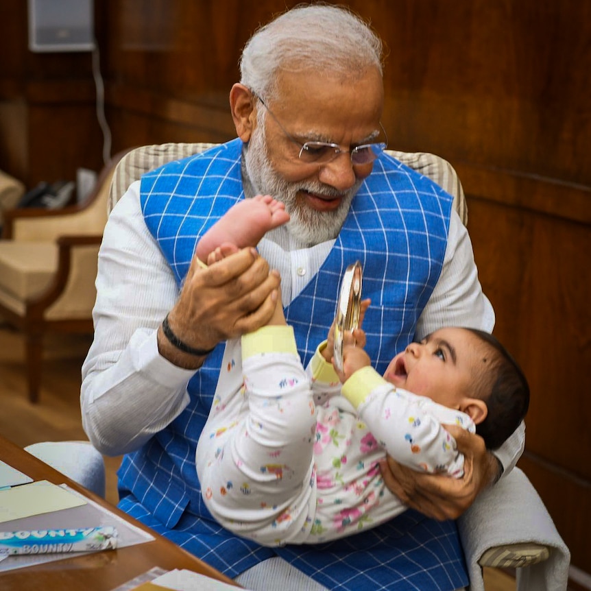 Narendra Modi, wearing a blue checked waistcoat, smiles as he plays with a baby on his lap, lifting its legs up