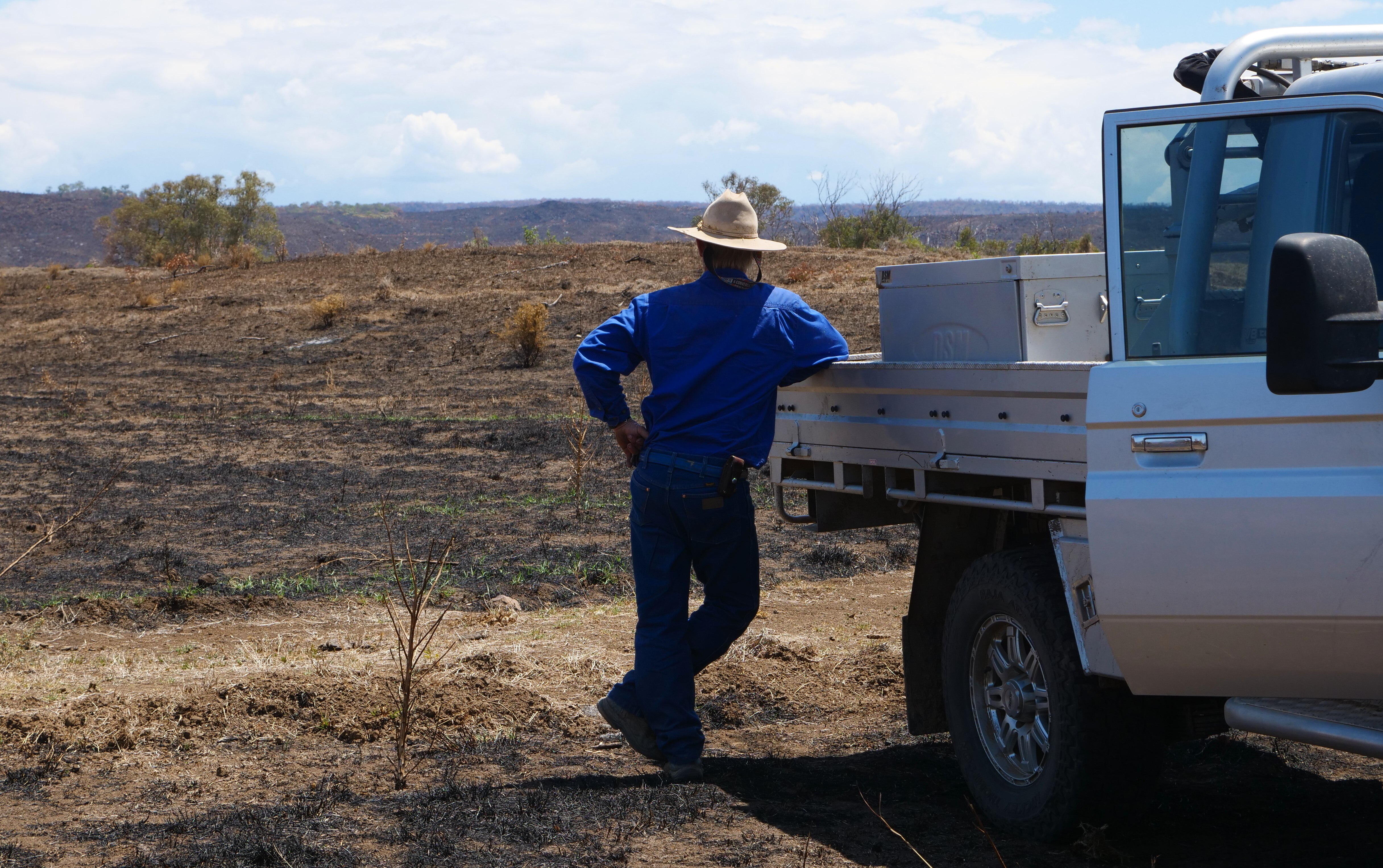 A middle aged man leans against a ute looking out over a completely blackened land.