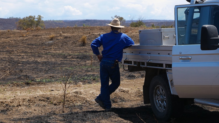 A middle aged man leans against a ute looking out over a completely blackened land.