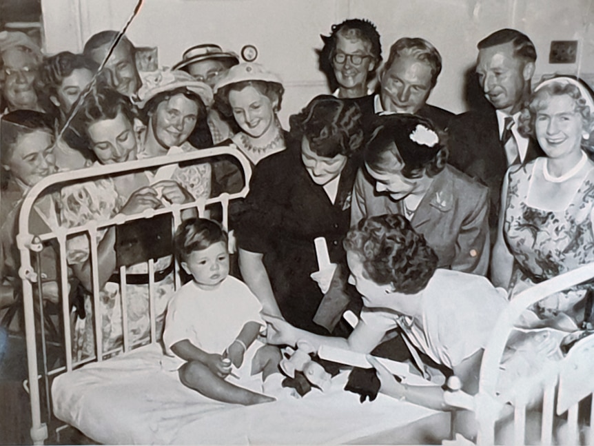 A black and white photo of a group of young women gathered around a cast iron hospital bed with a small child in it