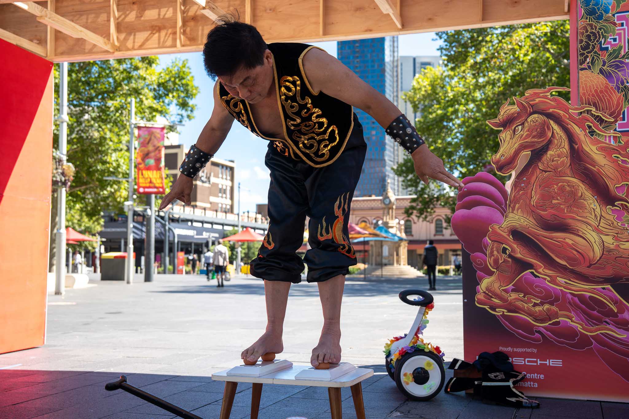 Mr Xiao balances on eggs on a small table while wearing a black costume with gold embroidery and flames on it outdoors.