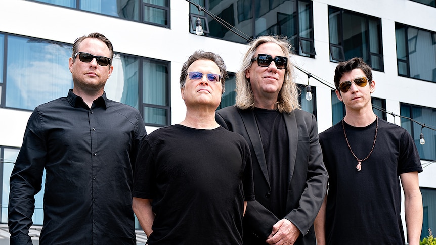 Four members of Violent Femmes, all dressed in black and wearing sunglasses, stand in front of a hotel