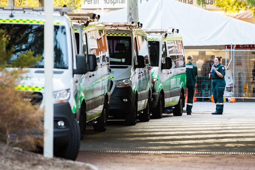 Two paramedics standing at the back of a row of ramped ambulances outside a hospital.
