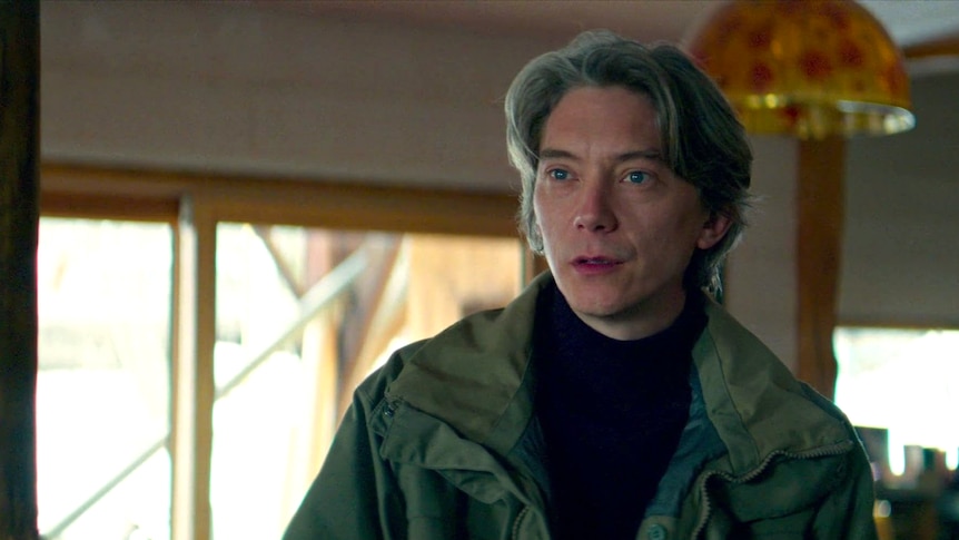 A man with blue eyes and floppy grey hair stands in a green jacket