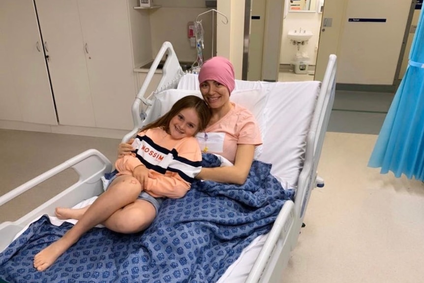 A woman in a beanie laying in a hospital bed, hugging a young girl to her chest.