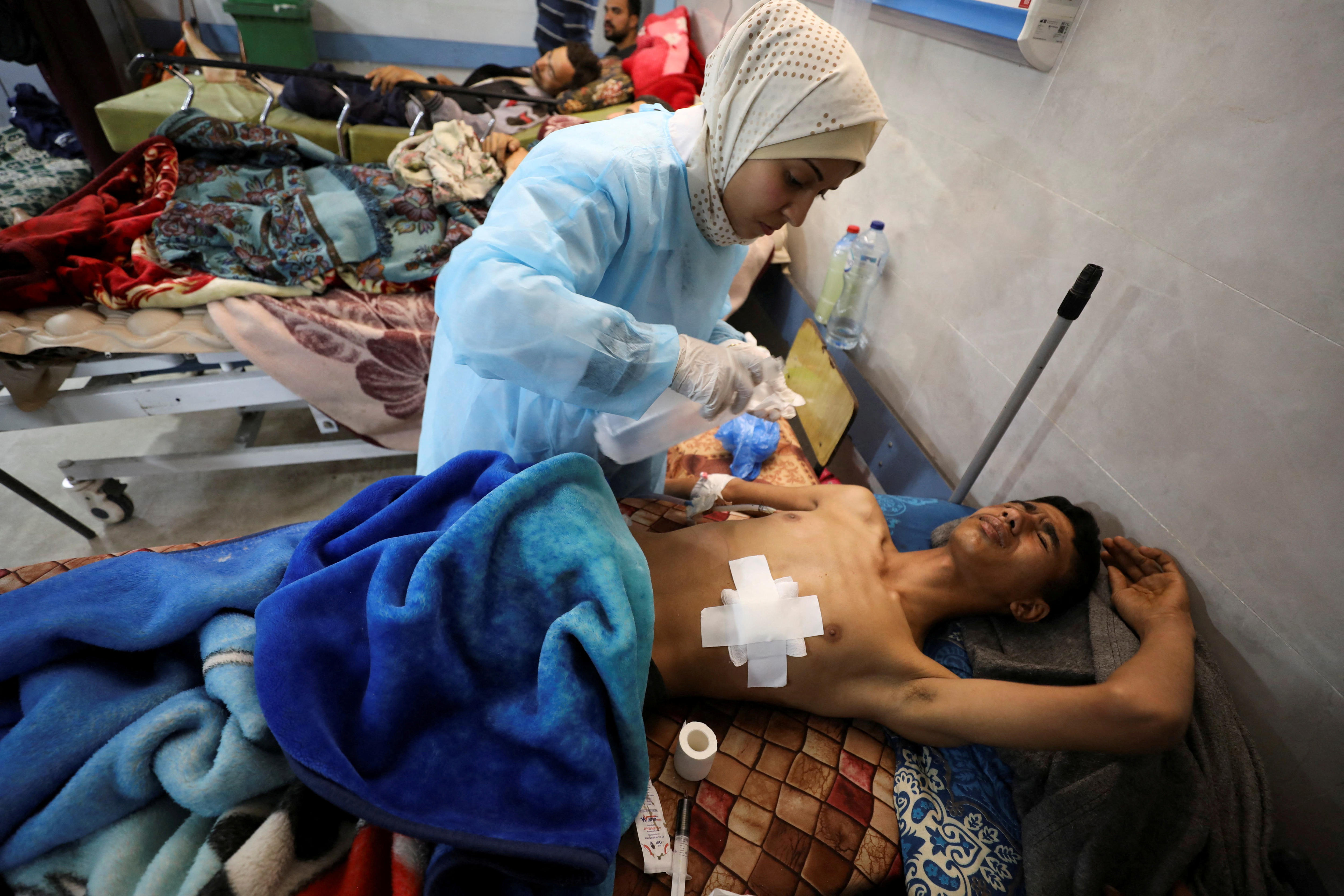 A medium photo of an injured man with a large bandage on his chest being treated by a woman in a crowded hospital.