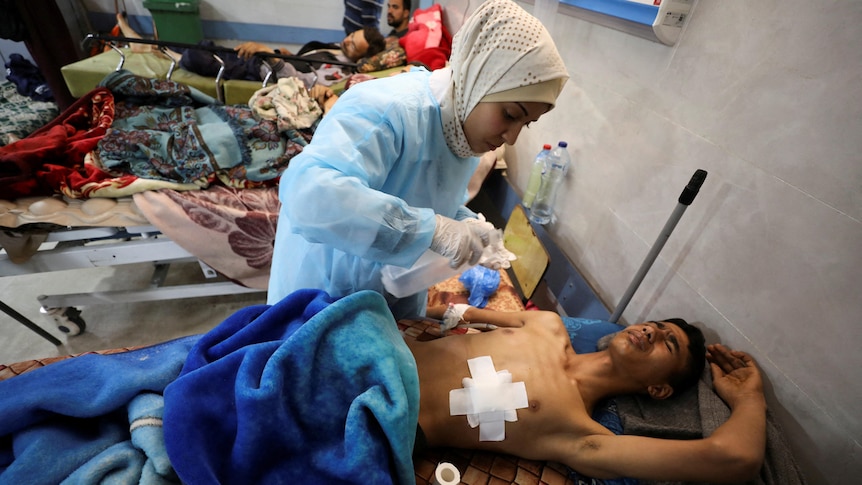 A medium photo of an injured man with a large bandage on his chest being treated by a woman in a crowded hospital.