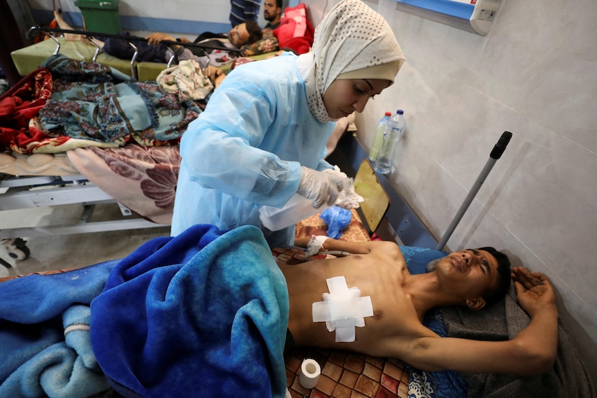 A medium photo of an injured man with a large bandage on his chest being treated by a woman in a crowded hospital.