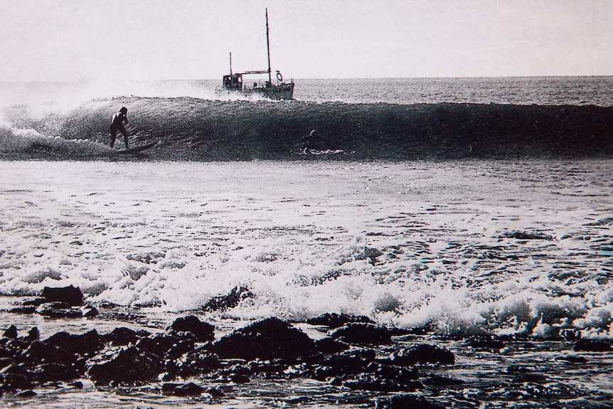 Black and white image of a surfer riding a long walled wave as a fishing boat approaches from behind