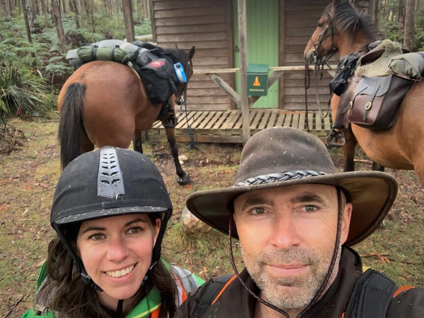 A woman and a man wearing horse riding gear taking a selfie in front of their two horses and an old hut.