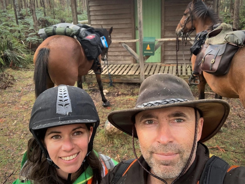 A woman and a man wearing horse riding gear taking a selfie in front of their two horses and an old hut.