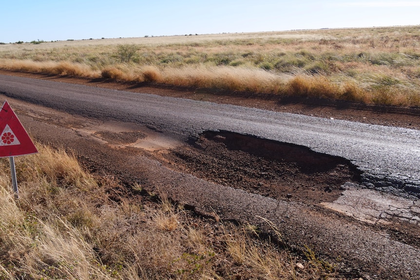 A pothole more than a meter long sits in a sealed road. A red warning sign is in the edge of the road.