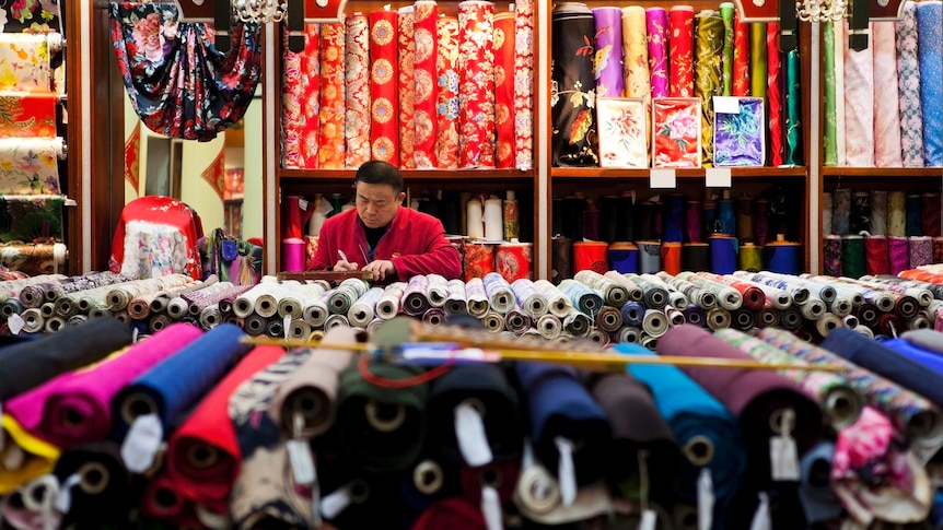 A silk seller sits in shop surrounded by many colourful fabric rolls