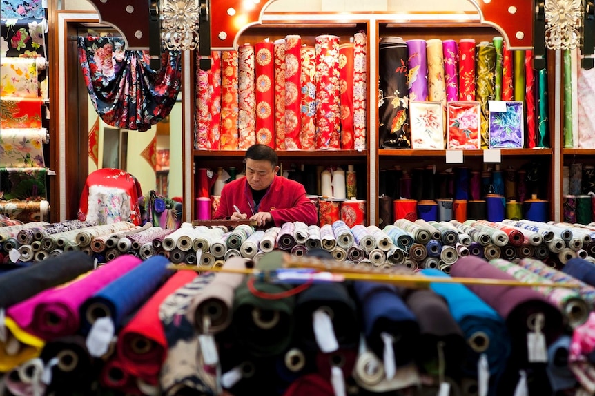A silk seller sits in shop surrounded by many colourful fabric rolls