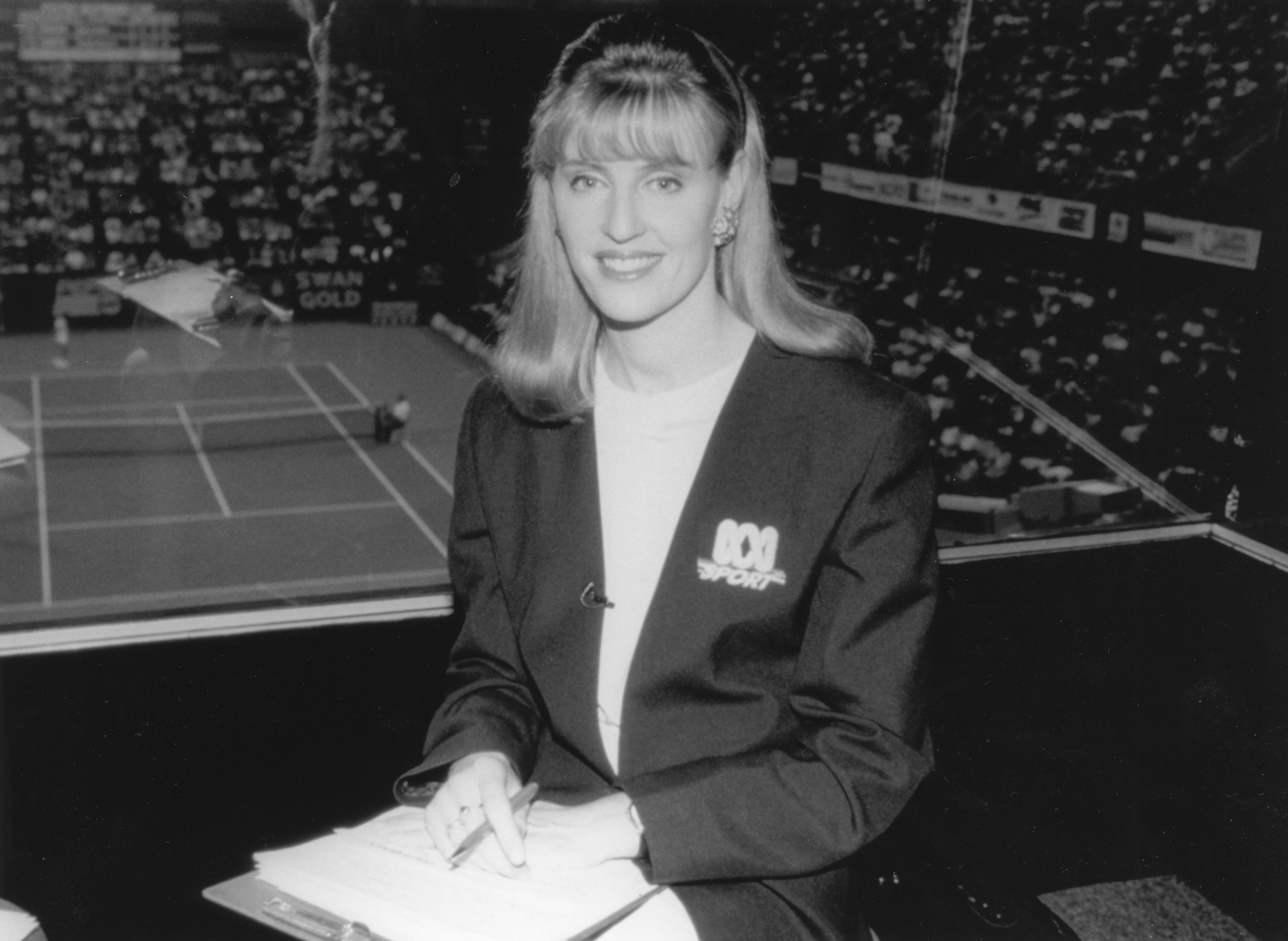 Black and white photo of Tighe in broadcast box with tennis court in background.