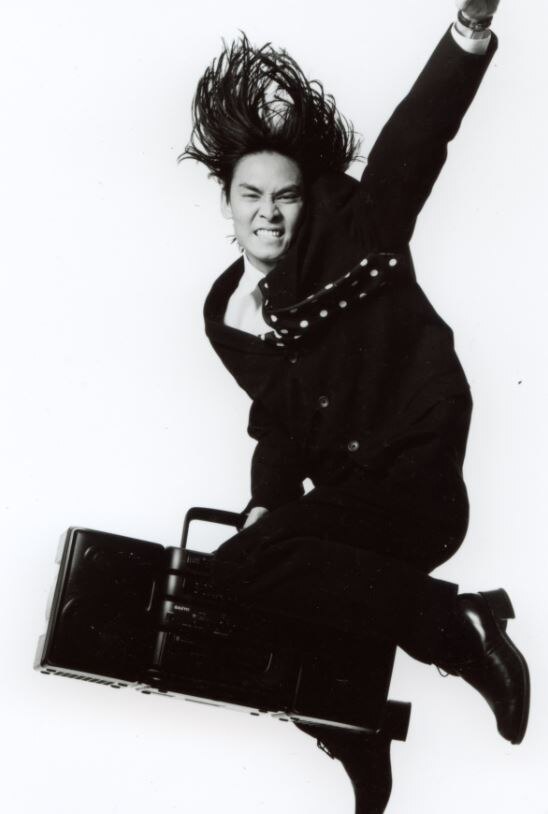 A man with long black hair, dressed in black, jumps in the air.