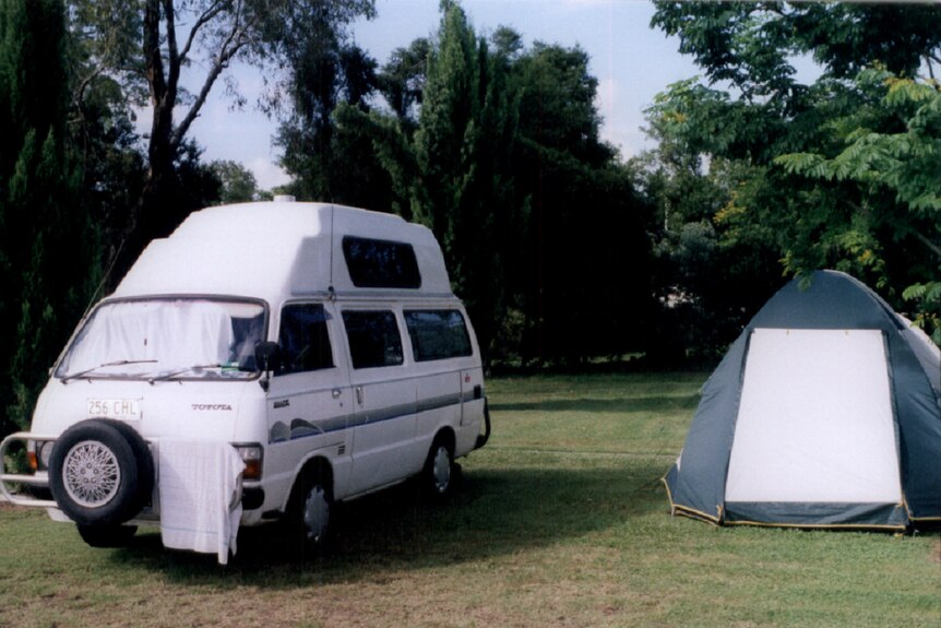 A white caravan parked next to a tent on grass with tall trees in the background.