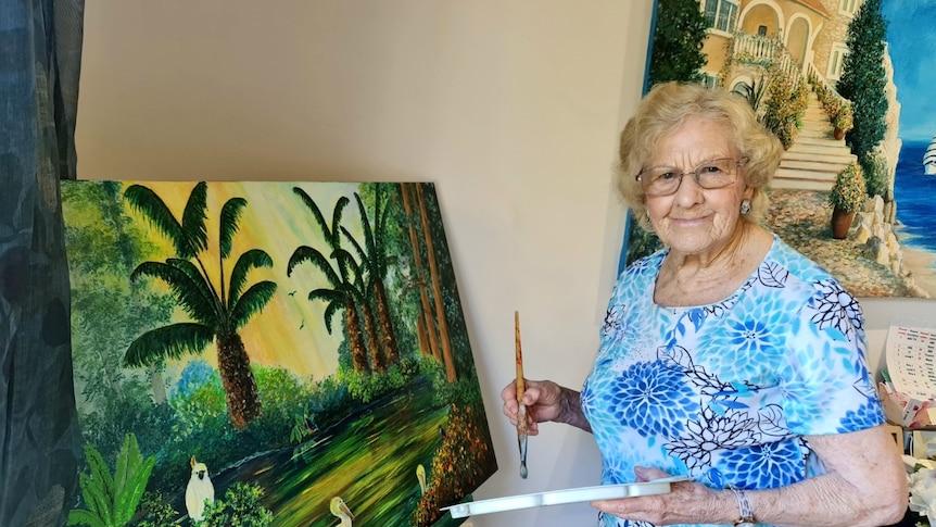 A smiling elderly woman wearing a blue patterned shirt, holding a paint brush and easel in front of a painting of a waterhole