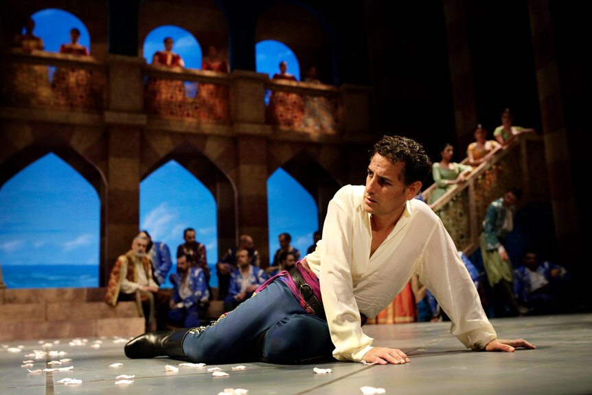 A man in an opera production lies on the floor in front of a chorus and some archways.