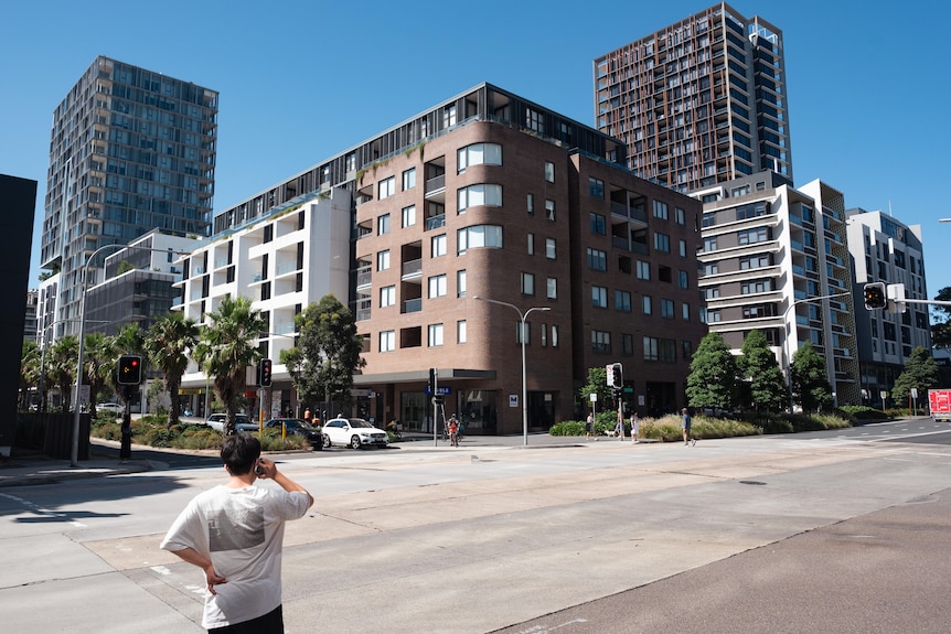 A man stands on the edge of a street in the foreground. Across the street are a number of large apartment buildings.