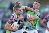 A Manly NRL player holds the ball with his right hand as he attempts to fend off a Canberra opponent.