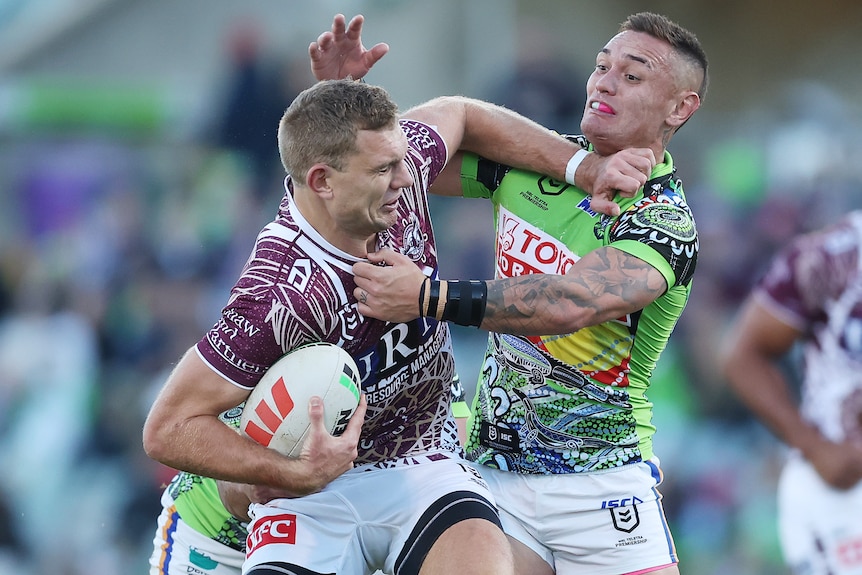 A Manly NRL player holds the ball with his right hand as he attempts to fend off a Canberra opponent.