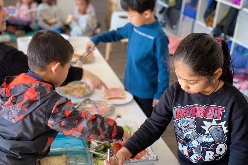 Children use tongs to serve themselves healthy food options from a table.